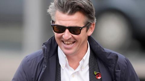 Joey Barton with grey hair wearing a navy coat, white shirt and navy checked suit with a poppy badge and black sunglasses. He is smiling.