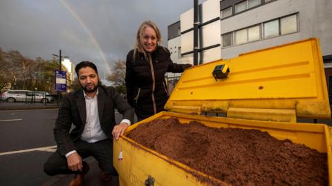 A man with dark hair and a beard is kneeling beside a full grit bin which is yellow and full of brown rock salt. he is wearing a white shirt and black suit. Beside him a woman with blond hair, wearing a dark coat, is stood smiling at the camera.