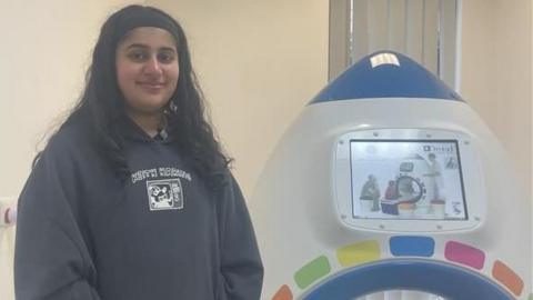 Manchester Royal patient, Manha is standing in front of the new rocket scanner device, she has long dark and is wearing a grey hoodie. 