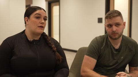 Parents Leah and Brandon Sanderson in a sit-down interview. Leah has brown hair in a plait and it wearing a brown top. Brandon has short brown hair, a short beard and is wearing a khaki green t-shirt with his hands clasped together on his lap.