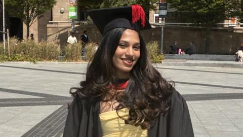 Person standing in a paved public square wearing a graduation gown and mortarboard with a tassel. A yellow dress is visible beneath the gown. Trees, ornamental grasses and stone buildings surround the square, with several people seated or walking in the background.