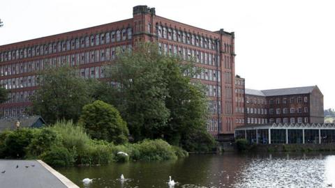 Red brick Belper North and East Mill behind trees with the River Derwent in front with swans gliding.