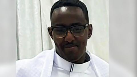 Mohamed Abdi Noor, a man with short dark hair and glasses, wearing white clothing and smiling.