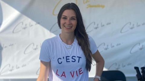 A woman with long brown hair is wearing a t-shirt that says 'C'est La Vie'.