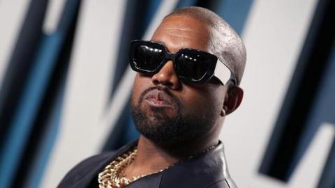 Kanye West with sunglasses on posing for a picture. He is also wearing a gold chain.