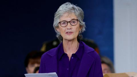 Catherine Connolly speaking. She is wearing silver earrings, glasses, a purple cardigan and a purple jacket.