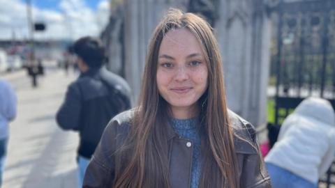 A young woman stands outdoors in daylight, facing the camera, with long hair and a jacket over a jumper. The background shows Parliament with passers‑by, railings, and a historic stone structure slightly out of focus.