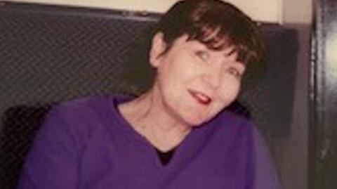 A woman wearing a purple jumper sits and looks into the camera.