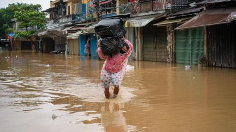A man in a raincoat wades through the floodwaters in Hoi An, carrying two large packs of items wrapped in black trash bags