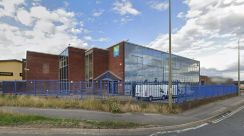 A general view picture of the depot building, which is made of brick and has a glass front. Three white vans are parked outside it.