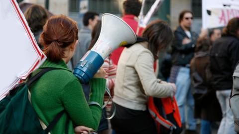 A woman wearing a green jumper facing away from the camera and holding a megaphone in crowd of people at protest, some holding placards.