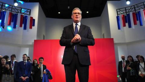 Keir Starmer wearing a dark-coloured suit and standing in front of a red background