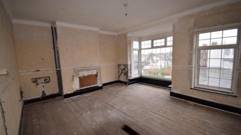 Inside one of the properties. There are uneven floorboards and bare walls, with the exception of some small areas where the remnants of some prevoius wallpaper can been seen peeling away.