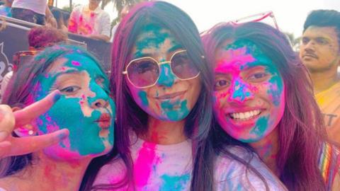 Three girls with long dark hair are covered in bright coloured powder, including magenta and turquoise, as part of the Holi festival of colour. They are all smiling. A man is behind them, covered with yellow powder.