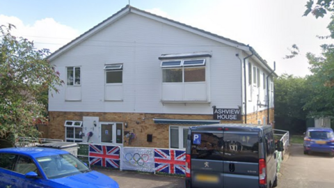 A Google street view image of Ashview House. It is a two-storey building. The top half is white, the bottom half is brick. There are Olympic and Union Jack flags hung outside it.