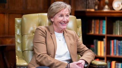 A close up shot of a middle-aged blonde woman sat in a leather chair in a room with a bookcase behind her smiling - Amanda Collier on The Traitors