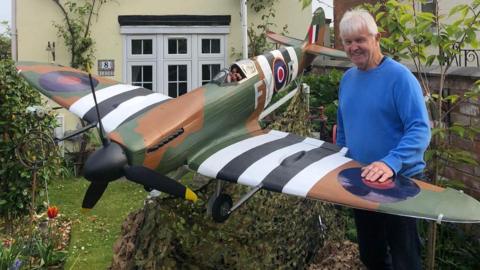 A large model Spitfire plane in a garden. The plane is dark green, white, black and brown with black propellers on its nose. It has an RAF circular logo on the side and camouflage netting underneath it. Standing next to it is Ray, who has white hair and is wearing a blue jumper. His hand is resting on the wing.