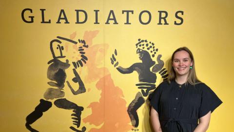 A woman in a black dress stands next to a yellow wall with black outlines of two men fighting with swords and shield with the word "Gladiators" written in black above them.