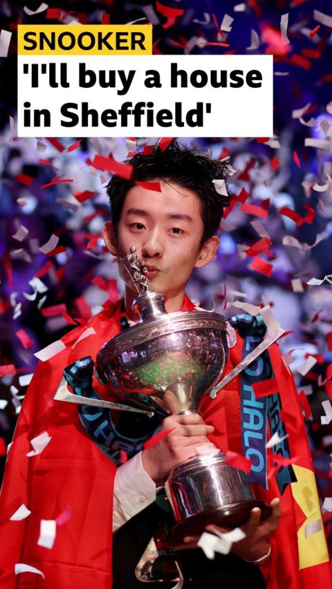 Wu Yize holding and kissing his World Snooker Championship Trophy. He is wearing the flag of China over his shoulders and there is red and white confetti falling.