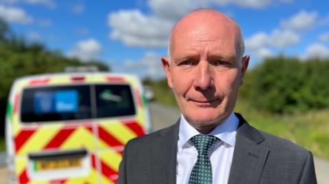 Darryl Preston who is wearing a grey suit, green tie and white shirt. He has no hair on the top of his head and short white hair on the sides. He is standing outside with a blurred out police van behind him on a road. Above him is blue sky with white clouds. 