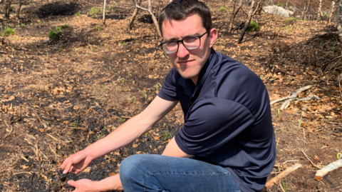 A man is kneeling on burnt ground and holding some blackened soil in his hands. He has brown hair and wears glasses with black frames. He is wearing a navy coloured t-shirt and blue jeans.