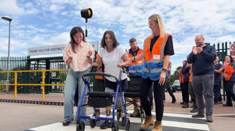 A woman wearing a white t-shirt is seen standing with the help of a stroller on wheels. She is surrounded by people who are cheering her on, as she uses a pelican crossing.