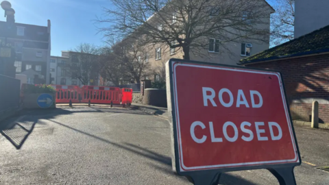 A red ROAD CLOSED sign in front of a fenced off part of a road surrounded by residential buildings.