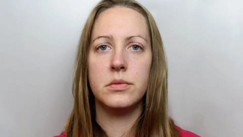 A mugshot of Lucy Letby. She has long light brown hair, is wearing a red top, and is staring into the camera lens.