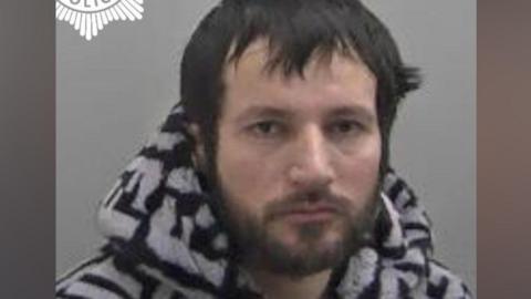 A police mugshot of a man wearing a black and white patterned jumper. He has black hair and a black beard. He is positioned against a light-coloured backdrop.