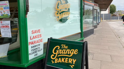 A shop window with a green circular logo, with yellow lettering that reads The Grange Bakery. Another sign in the window reads Save Lakes Parade. A green wooden sign sits outside the shop, with the same logo design, reading The Grange Bakery. 
