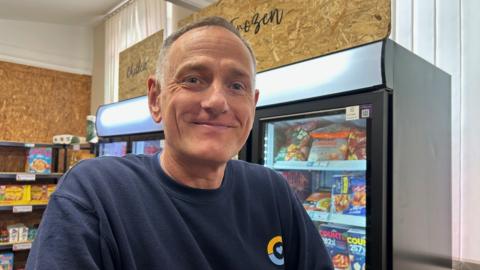 Simon Mitchell in the grocery. He is looking into the camera and smiling. He has short grey hair and is wearing a blue jumper. Behind him are large upright freezers filled with items of food, and shelves with packets of goods.