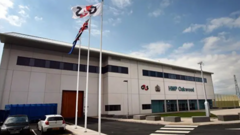 A two storey white building with the word HMP Oakwood in blue signage. There are three flag poles with a G4S flag, a Union Jack flag and blue flag while two cars, one black and another white, are parked outside the prison. 