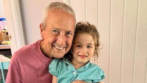Stan, who has short grey hair and wears a light red t-shirt, smiles as he holds Maggie while they pose for a picture. She is wearing a green and white checked summer school dress, and has her blonde wavy hair tied back in a ponytail. 