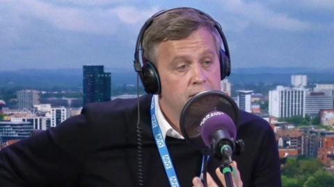James Roach wearing a black jumper on top of a white shirt and a blue NHS lanyard. He is wearing headphones and his face is partially obscured by a microphone and pop filter. He is in the studio and the studio backdrop shows an image of Southampton's skyline. 