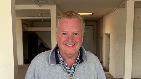 A man with greying red hair wearing a checked shirt and grey quarter-zip top standing in a derelict room. 