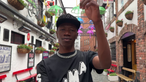 The Zambian rapper, Smack Jay, standing in an alleyway in Belfast. To his left and right are the entrances to two pubs. Above him are coloured lanterns hung across the alleyway.   