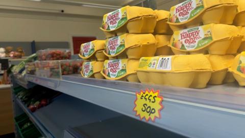 A stack of eggs in yellow boxes on a supermarket shelf. The packaging reads "the happy egg company", and below is a hand-written sticker that reads "Eggs 50 pence."