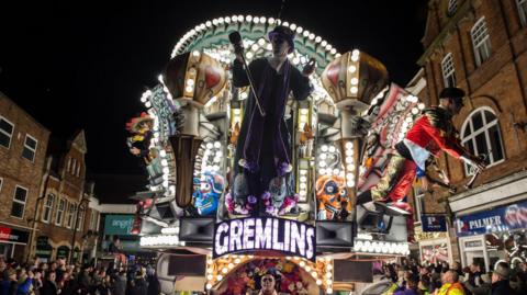 An illuminated carnival cart at night with many colours and lit-up sign saying 'Gremlins'.
There are lots of people surrounded the cart stood close to shops.