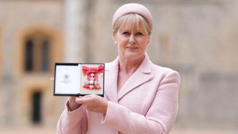 A mid-shot of Sarah Lancashire holding her CBE medal. She is wearing a pink beret and suit.
