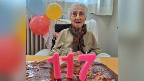 Maria Branyas, who has short grey hair, sat in front of a baked item with 117 candles on top and balloons behind her.