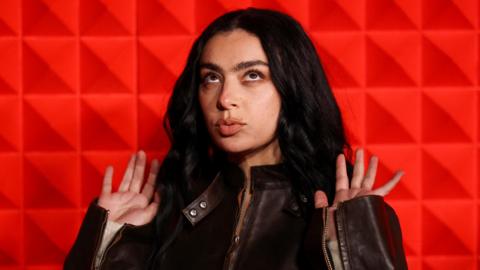 Charli XCX is looking up with her hands in a similar position. Her dark hair is slightly wavy and she has a slight pout.