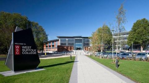 West Suffolk College sign on the left hand side of the image with the college building in the middle, a path leads up to the building with grass either side, cars are parked on the right hand side. 