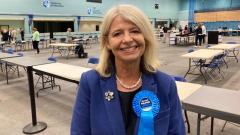 A woman with shoulder length blonde hair wearing a blue jacket and a blue rosette reading 'Harriett Baldwin' stands in a large hall with signs reading 'University of Worcester' on the wall.