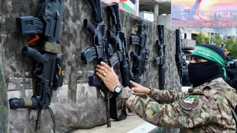 A Hamas soldier wearing a green Hamas headband lays rifles against a wall as they prepare for a hostage-prisoner swap with Israel in February 2025.