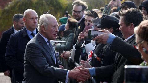 King Charles, wearing a grey suit and tie, shakes hands with people. The front of a large crowd can be seen to the right as the King stands on the left, with two men in suits behind him.