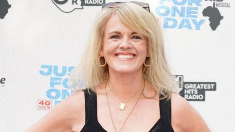 Sally Lindsay smiles at the camera. She has long blonde hair and wears gold hoop earrings two gold necklaces and a black sleeveless dress. 