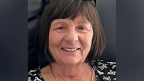 Margaret McGowan, a woman with dark finged bobbed hair, is seated on a black leather sofa and is wearing a black top with a white butterfly motif. She has black rimmed glasses on her head and a gold chain around her neck. 