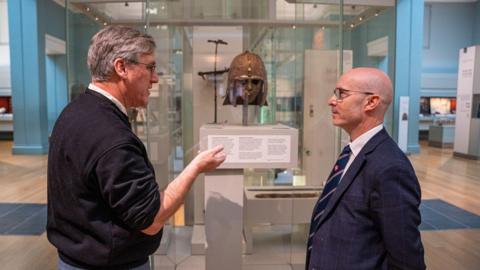 Two men talking to one another while stand in front of a decorative metal helmet in a museum case