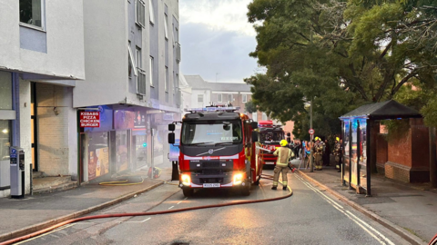 Fire engines parked outside the takeaway with thin smoke in the air outside the takeaways on the left, with flats above them.