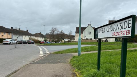 A sign saying Upper Churnside, Cirencester. There are houses in the distance with parked cars. There is a grazz verge on the right and road on the left.